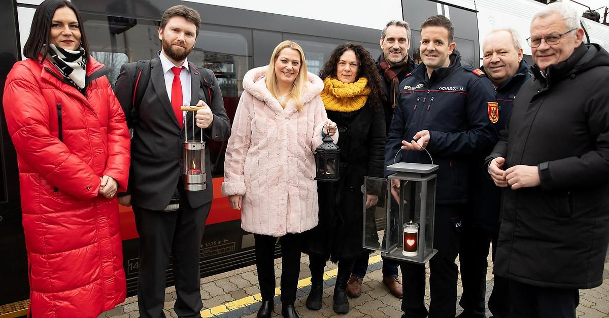 Fünf Personen stehen lächelnd auf einem Bahnsteig. Sie halten Laternen mit Kerzen darin. Ein Zug ist hinter ihnen.
