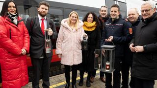 Fünf Personen stehen lächelnd auf einem Bahnsteig. Sie halten Laternen mit Kerzen darin. Ein Zug ist hinter ihnen.