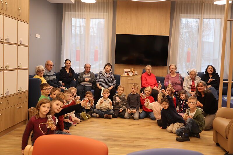 Eine Gruppe von Kindern und Erwachsenen versammelt sich in einem Raum und sitzt im Kreis, wobei sich Erwachsene auf Sofas hinter ihnen befinden. Alle halten kleine Geschenke in den Händen. Ein Fernseher ist an der Wand über einem Schrank montiert.
