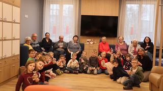 Eine Gruppe von Kindern und Erwachsenen versammelt sich in einem Raum und sitzt im Kreis, wobei sich Erwachsene auf Sofas hinter ihnen befinden. Alle halten kleine Geschenke in den Händen. Ein Fernseher ist an der Wand über einem Schrank montiert.