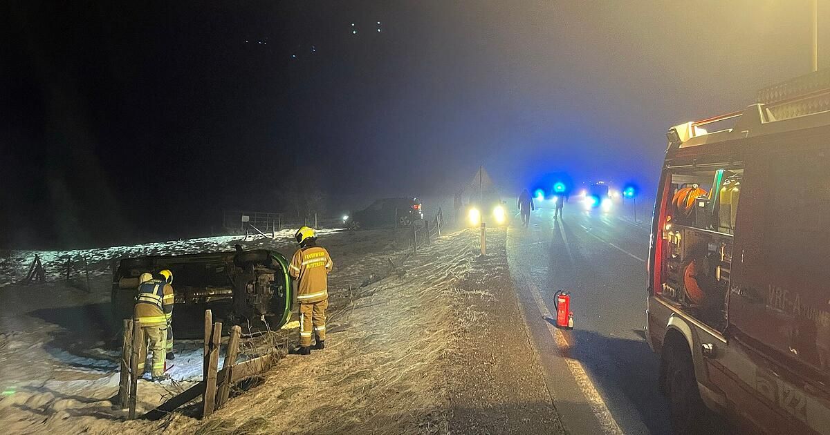 Feuerwehrleute kümmern sich um ein umgekipptes Auto auf einer nebligen Straße bei Nacht. Mehrere Notfallleuchten sind eingeschaltet.