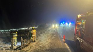 Feuerwehrleute kümmern sich um ein umgekipptes Auto auf einer nebligen Straße bei Nacht. Mehrere Notfallleuchten sind eingeschaltet.