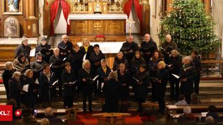 Ein Chor älterer Menschen, gekleidet in Schwarz und Weiß, singt in einer Kirche, mit einem Altar hinter ihnen. Der Altar ist mit einem goldenen Kreuz und Kerzen geschmückt. Ein Weihnachtsbaum steht auf der rechten Seite.