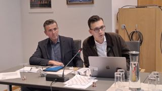 Zwei Männer in einem Besprechungsraum. Einer mit Laptop und der andere mit Tablet. Beide tragen Brillen. Ein Mikrofon steht vor ihnen.