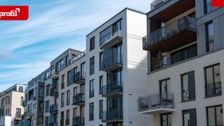 Ein modernes Wohngebäude mit weißer Außenfassade, mehreren Stockwerken und Balkonen. Jede Etage hat einen Balkon mit Geländer, und einige haben Topfpflanzen. Der Himmel ist klar mit einigen Wolken.