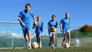 Vier Kinder in blauen Trikots rennen über ein Fußballfeld mit Bergen im Hintergrund. Die Kinder halten Fußballbälle. Das Feld ist eingezäunt und hat ein Tornetz.
