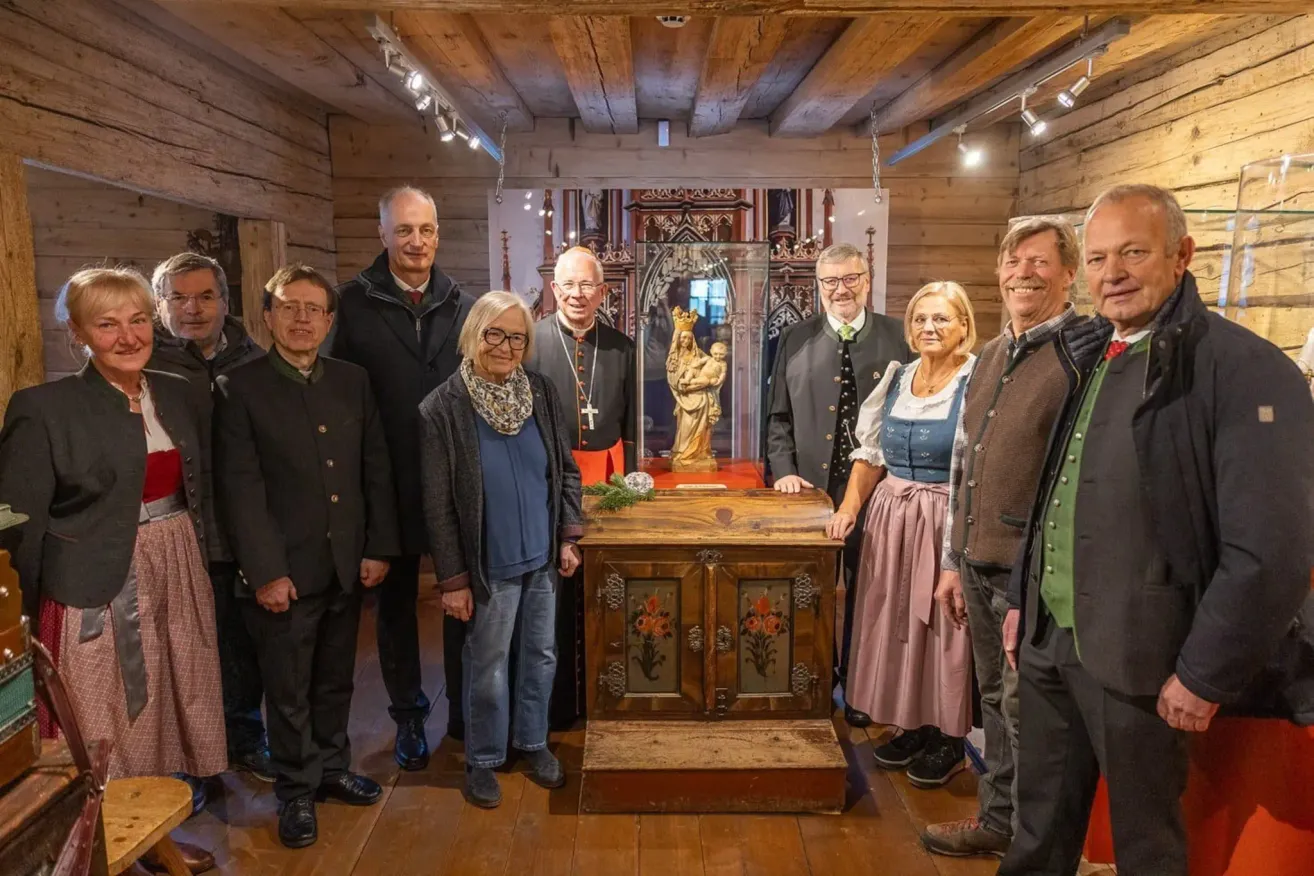 Eine Gruppe von Menschen, darunter Männer und Frauen, steht um einen hölzernen Altar herum. Hinter ihnen steht eine Statue eines Heiligen und eine Glasvitrine.