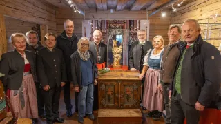 Eine Gruppe von Menschen, darunter Männer und Frauen, steht um einen hölzernen Altar herum. Hinter ihnen steht eine Statue eines Heiligen und eine Glasvitrine.