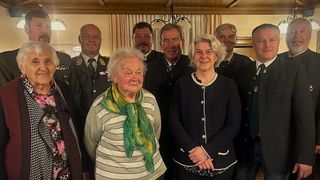 Eine Gruppe von sieben Personen, darunter zwei Frauen, eine ältere und eine jüngere, steht gemeinsam in einem Raum und posiert für ein Foto.