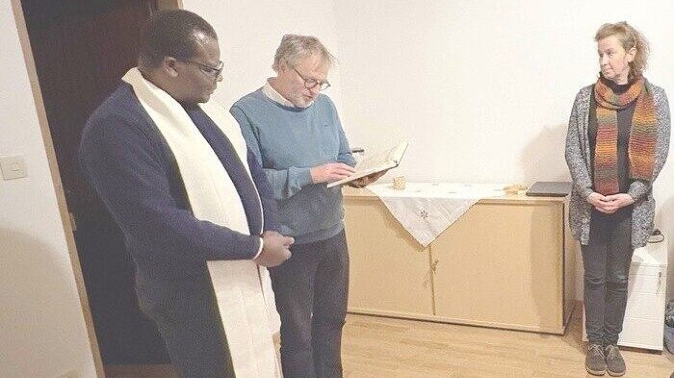 Zwei Männer stehen in einem Raum, einer trägt eine weiße Stola und eine Brille, der andere einen blauen Pullover und eine Brille und liest ein Buch. Hinter ihnen steht ein Tisch mit einem weißen Tuch.