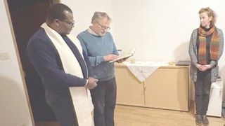 Zwei Männer stehen in einem Raum, einer trägt eine weiße Stola und eine Brille, der andere einen blauen Pullover und eine Brille und liest ein Buch. Hinter ihnen steht ein Tisch mit einem weißen Tuch.