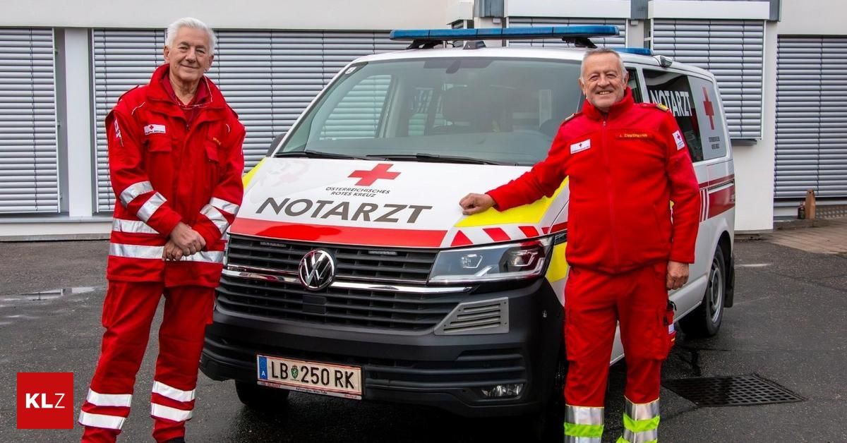 Zwei Männer in roten Uniformen stehen neben einem weißen Krankenwagen mit dem Wort 'NOTARZT' an der Seite. Der Mann auf der linken Seite trägt eine Halskette.