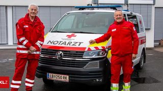 Zwei Männer in roten Uniformen stehen neben einem weißen Krankenwagen mit dem Wort 'NOTARZT' an der Seite. Der Mann auf der linken Seite trägt eine Halskette.