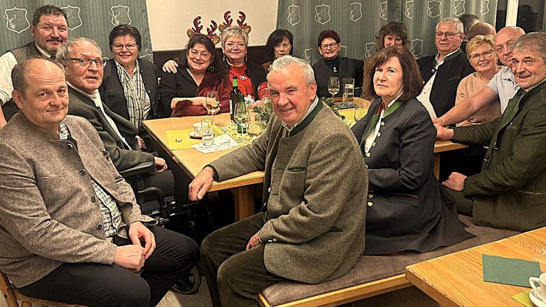 Eine Gruppe von Menschen, darunter ein Mann und Frauen, sitzt um einen Tisch herum, wahrscheinlich in einem Restaurant, lächelt und posiert für ein Foto. Sie tragen Winterkleidung und halten Weingläser in der Hand. Auf dem Tisch stehen Weinflaschen, Gläser und Tassen. Einige Leute tragen eine Brille. Hinter ihnen befindet sich eine Wand mit Weihnachtsdekoration und einem Vorhang.