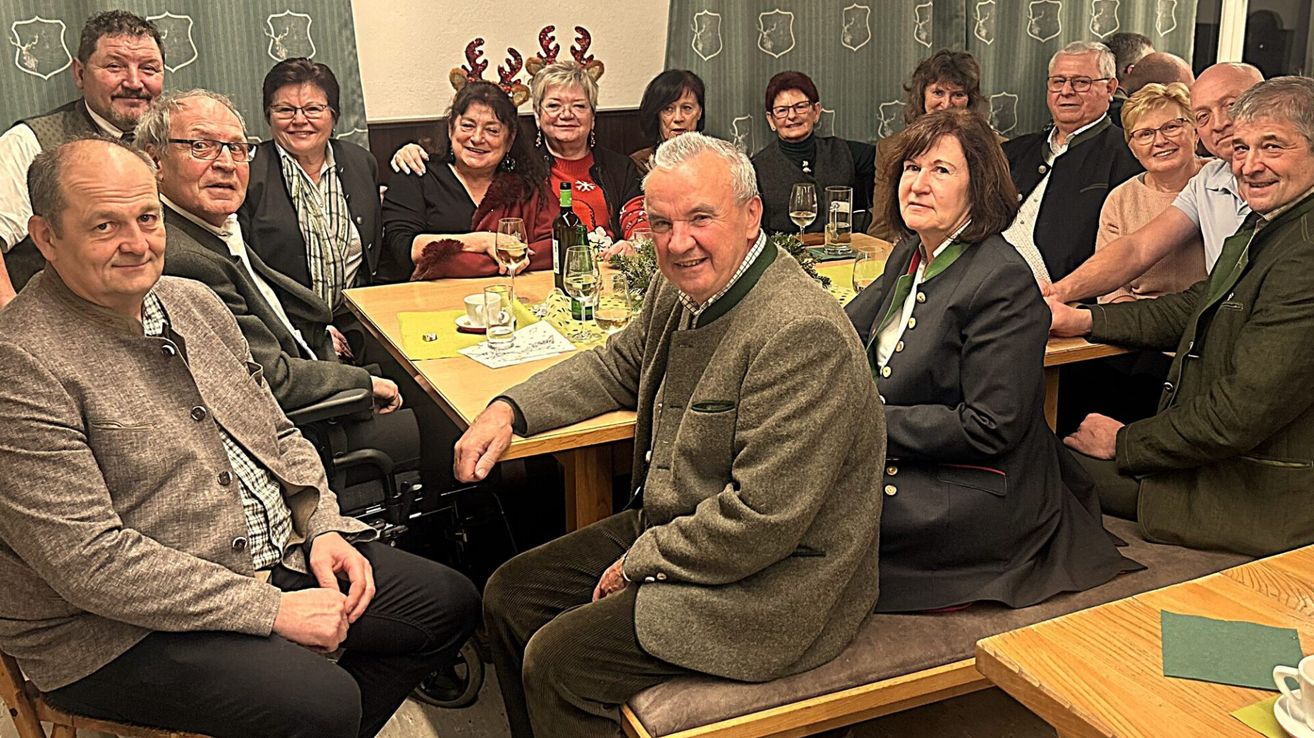 Eine Gruppe von Menschen, darunter ein Mann und Frauen, sitzt um einen Tisch herum, wahrscheinlich in einem Restaurant, lächelt und posiert für ein Foto. Sie tragen Winterkleidung und halten Weingläser in der Hand. Auf dem Tisch stehen Weinflaschen, Gläser und Tassen. Einige Leute tragen eine Brille. Hinter ihnen befindet sich eine Wand mit Weihnachtsdekoration und einem Vorhang.