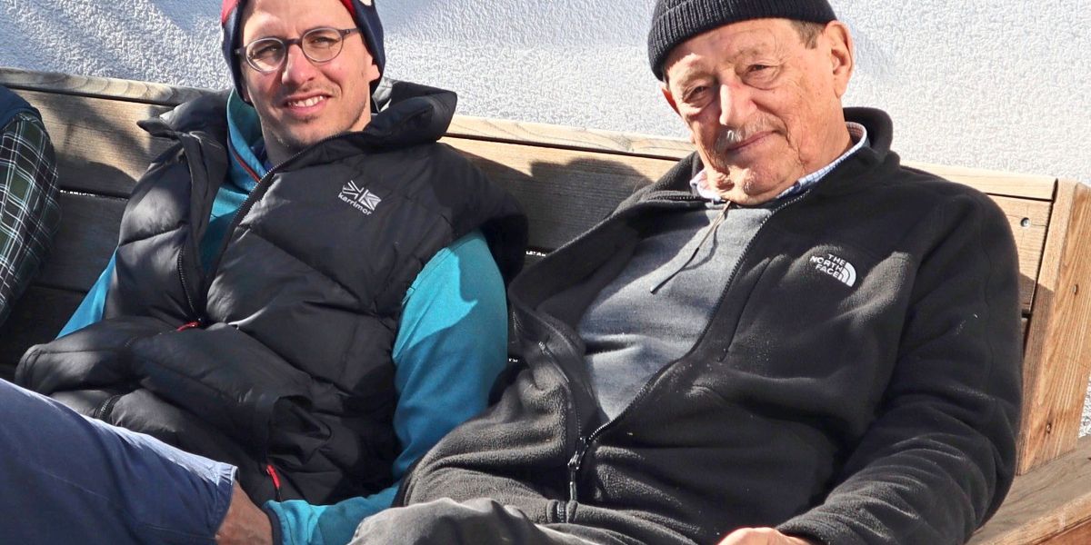 Zwei Männer sitzen auf einer Holzbank und lächeln für ein Foto. Der linke Mann trägt eine schwarze und blaue Jacke mit einem weißen Logo. Der rechte Mann trägt eine schwarze Jacke und eine schwarze Mütze.