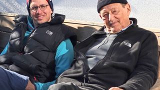 Zwei Männer sitzen auf einer Holzbank und lächeln für ein Foto. Der linke Mann trägt eine schwarze und blaue Jacke mit einem weißen Logo. Der rechte Mann trägt eine schwarze Jacke und eine schwarze Mütze.