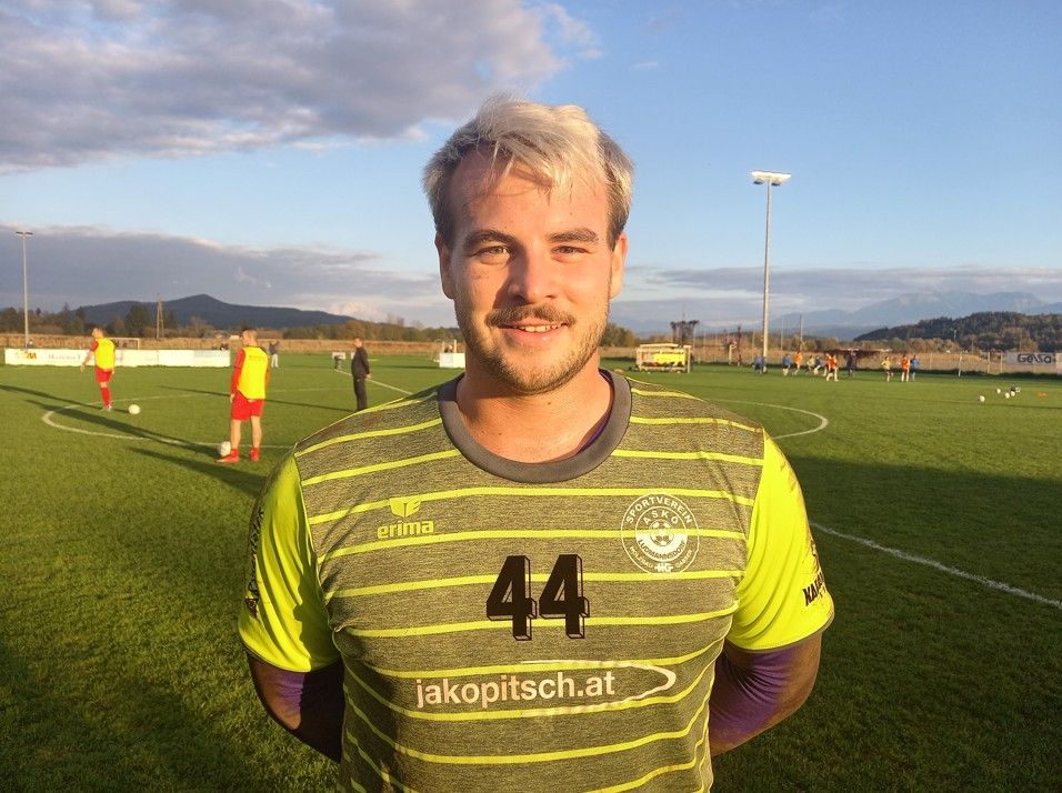 Ein Mann in einem Fußballtrikot steht lächelnd auf einem Fußballfeld. Das Feld ist grün mit einer weißen Linie und einem Fußballtor. Im Hintergrund üben Spieler.