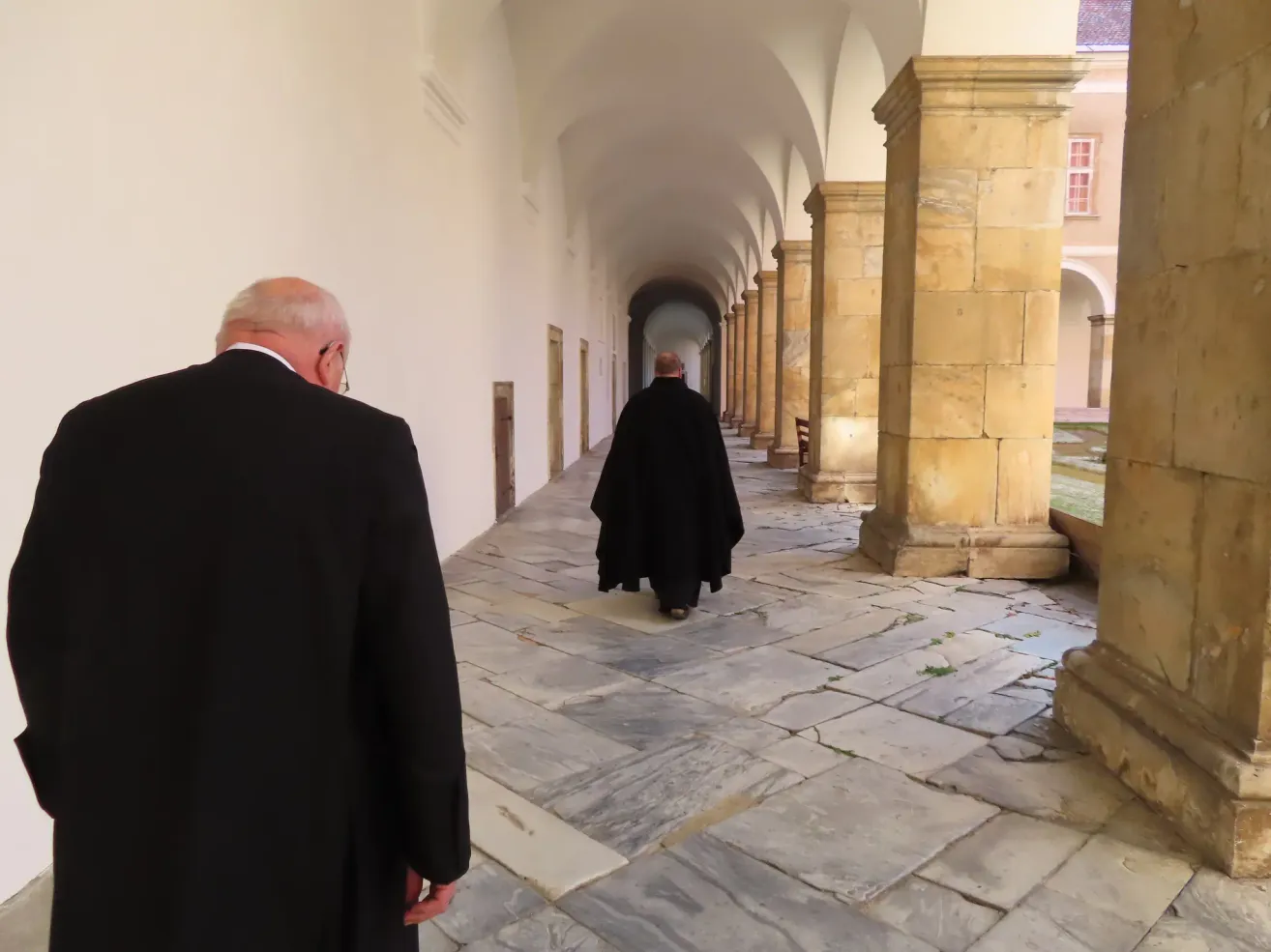 Zwei Männer in schwarzen Mänteln gehen einen Steinweg in einem alten Gebäude mit gewölbten Decken und Säulen entlang.