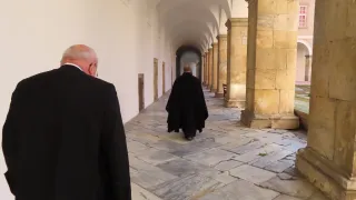 Zwei Männer in schwarzen Mänteln gehen einen Steinweg in einem alten Gebäude mit gewölbten Decken und Säulen entlang.