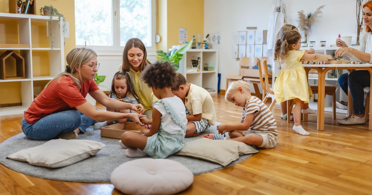 Mehrere Kinder spielen auf dem Boden, angeleitet von zwei Erwachsenen. Sie sind in eine spielerische Aktivität vertieft, umgeben von Spielzeug und Kissen. Der Raum hat Holzböden und ist von natürlichem Licht durchflutet.