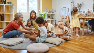 Mehrere Kinder spielen auf dem Boden, angeleitet von zwei Erwachsenen. Sie sind in eine spielerische Aktivität vertieft, umgeben von Spielzeug und Kissen. Der Raum hat Holzböden und ist von natürlichem Licht durchflutet.