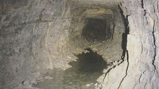 Ein unterirdischer Tunnel mit fließendem Wasser, Wände aus Stein mit Rissen und Schutt.