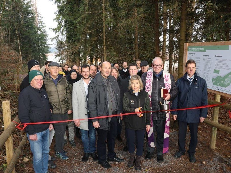 Eine Gruppe von Menschen versammelt sich in einem Wald, hinter einem roten Band stehend. Eine Frau schneidet das Band, während ein Mann ein Buch in der Hand hält. Hinter ihnen steht ein Schild mit Text.