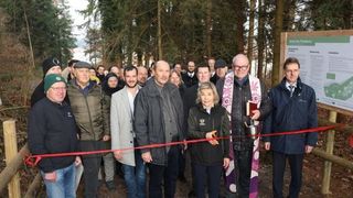 Eine Gruppe von Menschen versammelt sich in einem Wald, hinter einem roten Band stehend. Eine Frau schneidet das Band, während ein Mann ein Buch in der Hand hält. Hinter ihnen steht ein Schild mit Text.