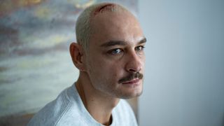 Ein kahlköpfiger Mann mit einer Narbe auf der Stirn, der ein graues T-Shirt trägt, starrt den Betrachter ernsthaft an.