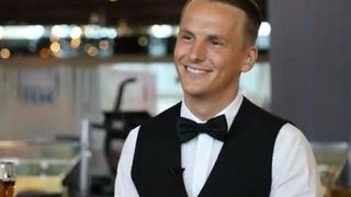 Ein Mann lächelt und posiert für ein Foto. Er trägt eine schwarze Weste und ein weißes Hemd. Im Hintergrund ist ein unscharfer Hintergrund zu sehen.