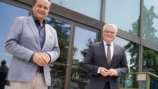 Zwei Männer in Anzügen stehen vor einem Gebäude mit Glastüren. Einer lächelt, der andere trägt eine Brille. Ein Tisch mit Flaschen ist durch das Glas zu sehen.