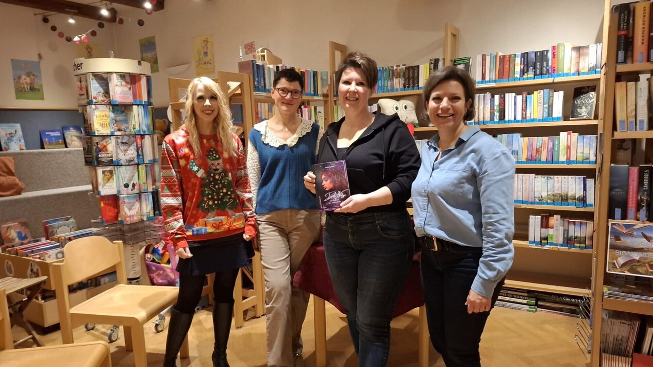 Vier Frauen stehen in einer Buchhandlung mit Regalen voller Bücher. Die Frau in der Mitte hält ein Buch mit dem Titel 'Dahlia'. Die Frau links trägt einen Weihnachtspullover.
