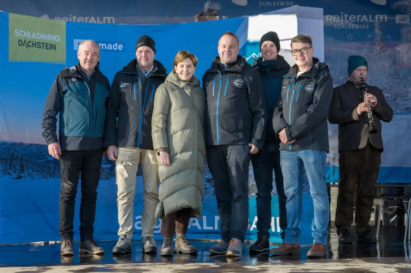 Sechs Personen stehen in einer Reihe und tragen Winterkleidung, wahrscheinlich für ein Foto. Sie alle lächeln. Hinter ihnen befindet sich ein blauer Banner mit weißem Text.