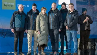 Sechs Personen stehen in einer Reihe und tragen Winterkleidung, wahrscheinlich für ein Foto. Sie alle lächeln. Hinter ihnen befindet sich ein blauer Banner mit weißem Text.