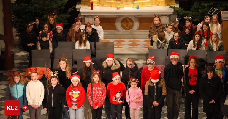 Ein Kinderchor in Weihnachtskleidung steht vor einem Altar in einer Kirche und singt mit Mikrofonen. Einige tragen Weihnachtsmützen.