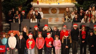 Ein Kinderchor in Weihnachtskleidung steht vor einem Altar in einer Kirche und singt mit Mikrofonen. Einige tragen Weihnachtsmützen.