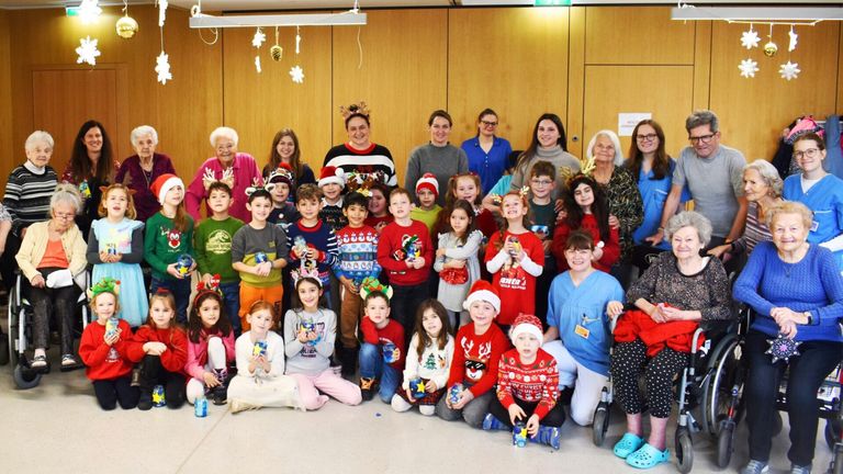 Eine Gruppe von Erwachsenen und Kindern steht in einem Raum, lächelt und hält Weihnachtsgeschenke. Die Erwachsenen sind in festlichen Pullovern gekleidet, während die Kinder Weihnachtsmützen tragen. Sie scheinen für ein Foto zu posieren.
