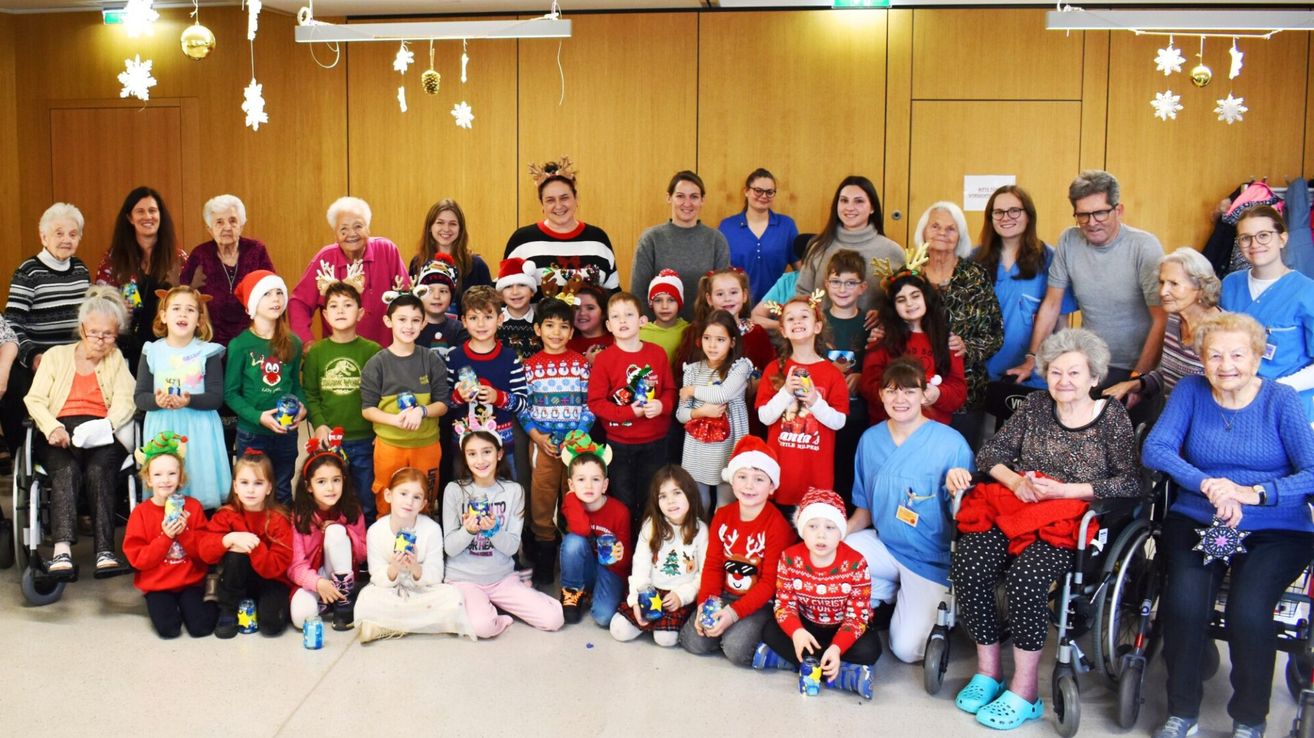 Eine Gruppe von Erwachsenen und Kindern steht in einem Raum, lächelt und hält Weihnachtsgeschenke. Die Erwachsenen sind in festlichen Pullovern gekleidet, während die Kinder Weihnachtsmützen tragen. Sie scheinen für ein Foto zu posieren.