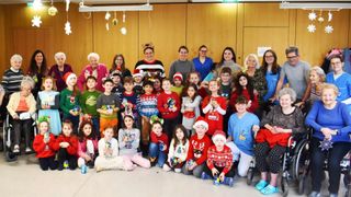 Eine Gruppe von Erwachsenen und Kindern steht in einem Raum, lächelt und hält Weihnachtsgeschenke. Die Erwachsenen sind in festlichen Pullovern gekleidet, während die Kinder Weihnachtsmützen tragen. Sie scheinen für ein Foto zu posieren.