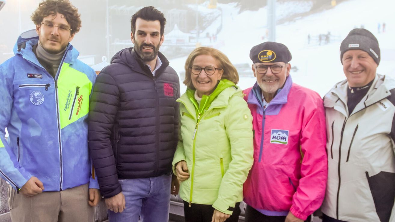 Drei Menschen stehen vor einem verschneierten Hintergrund. Der Mann links trägt eine dunkle Jacke und Jeans. Die Frau in der Mitte trägt eine grüne Jacke und eine Brille. Der Mann rechts trägt eine rosa Jacke mit Logo.