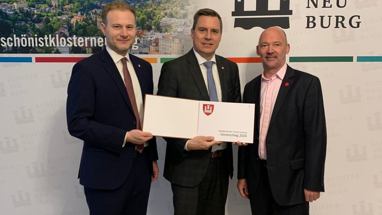 Drei Männer in formeller Kleidung, einer hält ein gerahmtes Zertifikat, stehen vor einer Kulisse mit einer Stadtlandschaft und einem Logo.