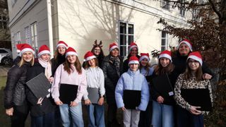 Eine Gruppe von Menschen, sowohl Erwachsene als auch Kinder, steht vor einem Gebäude, alle tragen Weihnachtsmützen und halten Bücher in der Hand und lächeln für ein Foto.