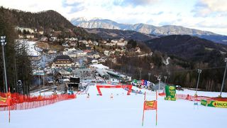 Luftaufnahme eines Skigebiets mit einem verschneiten Berg im Hintergrund. Die Pisten sind mit roten Fahnen markiert, und es sind Skifahrer zu sehen.