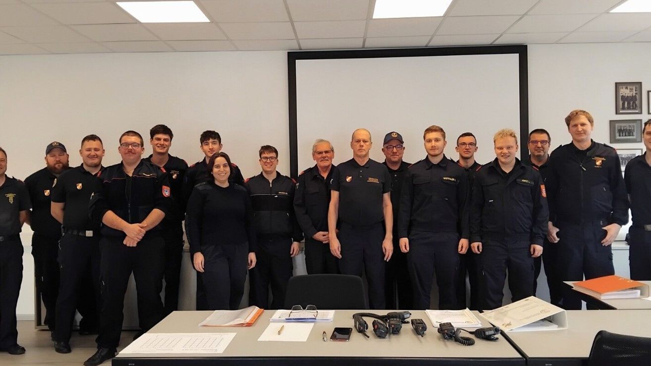 Eine Gruppe von Feuerwehrleuten in Uniform steht hinter einem Tisch mit verschiedenen Geräten. Eine Frau ist unter ihnen. Sie befinden sich in einem Raum mit einer Tafel und weißen Wänden.