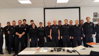 Eine Gruppe von Feuerwehrleuten in Uniform steht hinter einem Tisch mit verschiedenen Geräten. Eine Frau ist unter ihnen. Sie befinden sich in einem Raum mit einer Tafel und weißen Wänden.