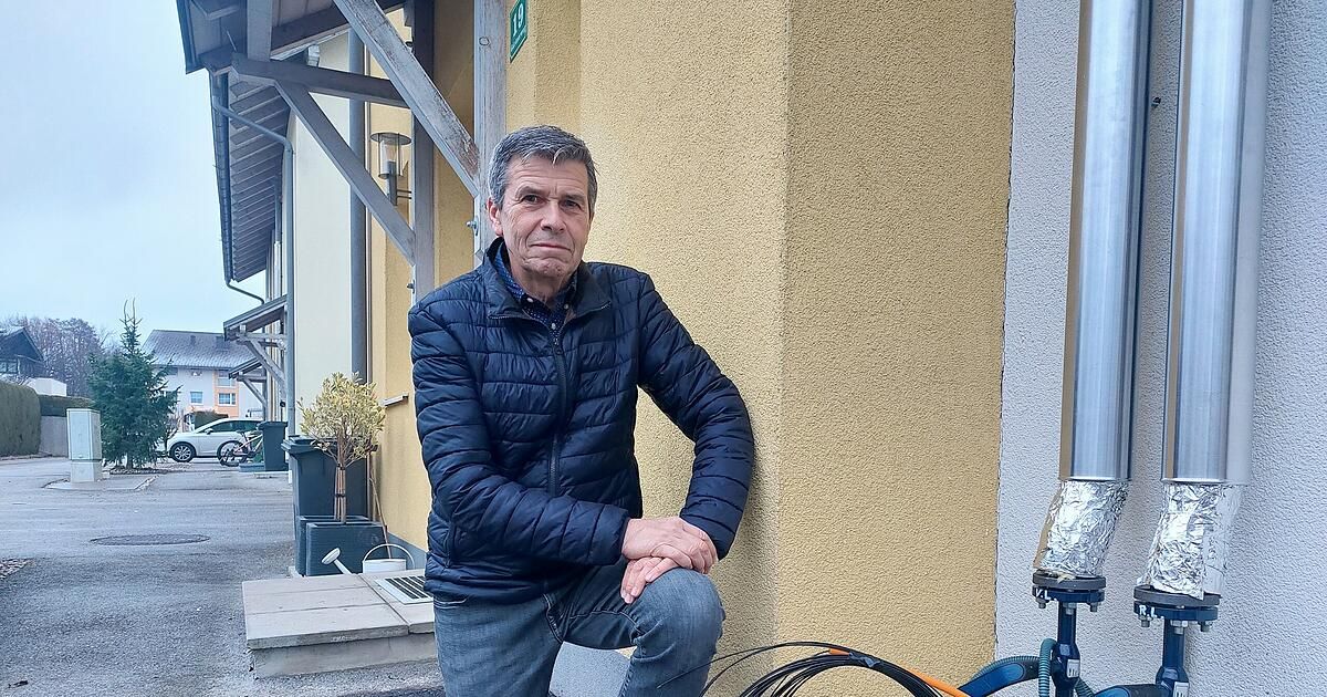 Ein älterer Mann in einer blauen Jacke und Jeans sitzt auf einer Mauer vor einem Gebäude, die Hände auf den Knien.