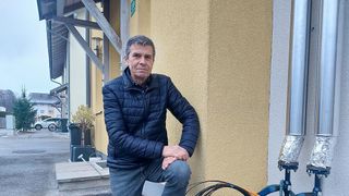 Ein älterer Mann in einer blauen Jacke und Jeans sitzt auf einer Mauer vor einem Gebäude, die Hände auf den Knien.