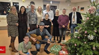 Eine Gruppe von Menschen posiert für ein Bild in einem weihnachtlich geschmückten Raum, einer spielt Gitarre und einer trägt einen Hut. Geschenke und ein Weihnachtsbaum sind vorhanden.