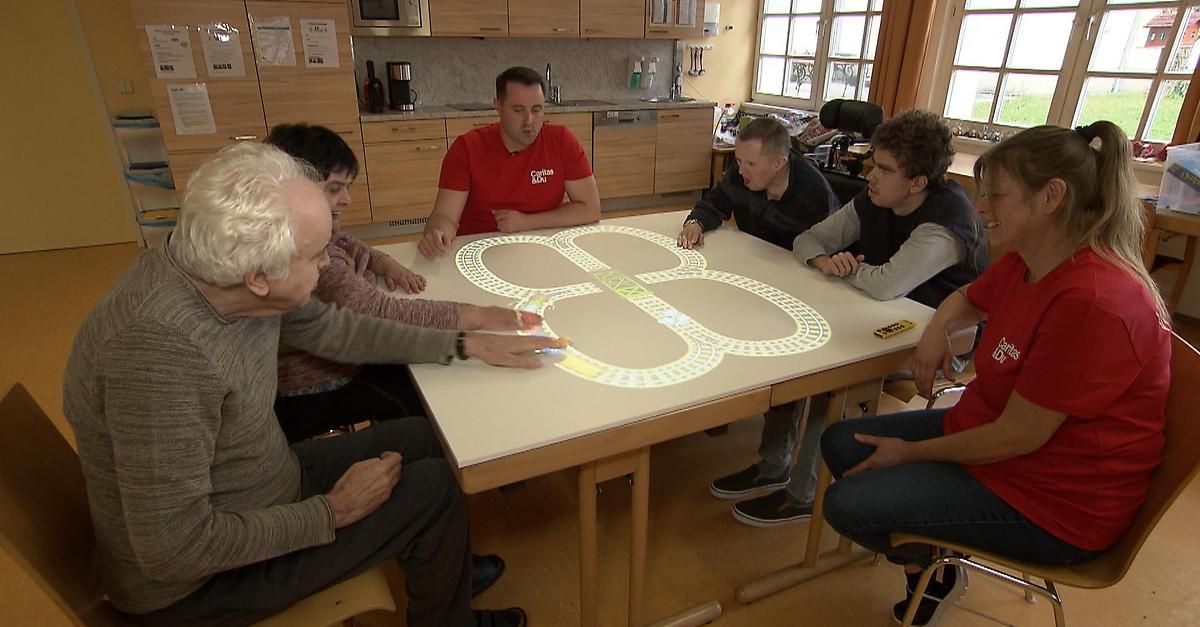 Fünf Personen sitzen um einen Tisch herum und konzentrieren sich auf ein großes beleuchtetes Design einer Eisenbahnstrecke auf dem Tisch. Eine Person zeigt auf das Design.